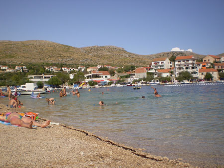 Belebter Kiesstrand mit Badenden und flachem, klarem Wasser.